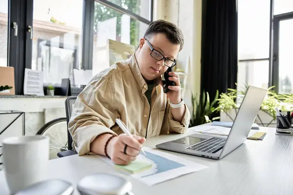 Un joven con síndrome de Down toma notas mientras está en una llamada telefónica en un entorno de oficina moderno. - foto de stock