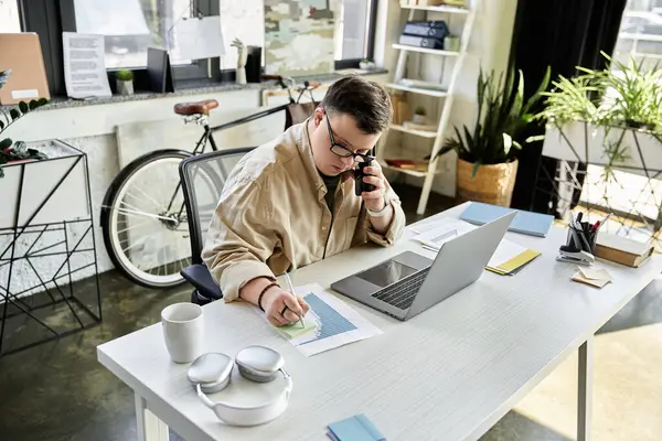 A young man with Down syndrome is on the phone at his desk, working on a graph. — Stock Photo