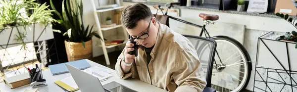 A young man with Down syndrome sits at his desk, taking a phone call while working on his laptop. — Stock Photo