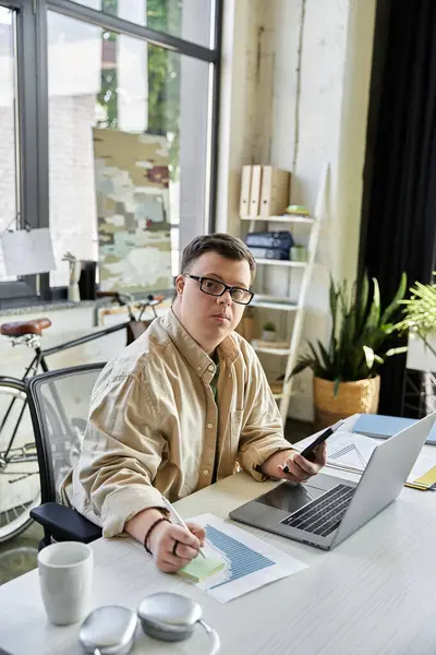 A young man with Down syndrome works on his laptop and a sticky note, looking focused and determined. — Stock Photo