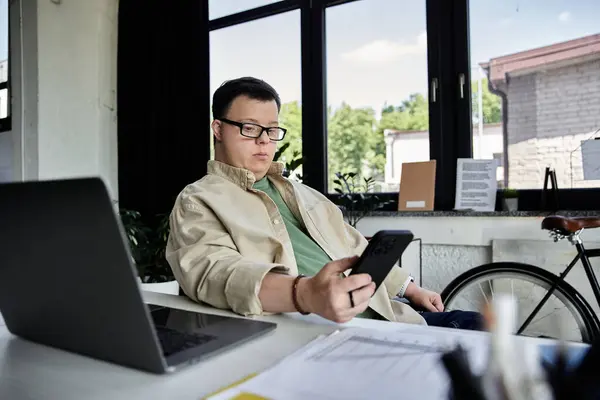 A young man with Down syndrome is sitting at a desk and checking his phone. — Stock Photo