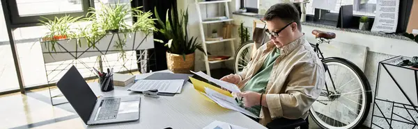 A young man with Down syndrome sits at a desk reviewing documents, while a bicycle rests against the wall behind him. — Stock Photo