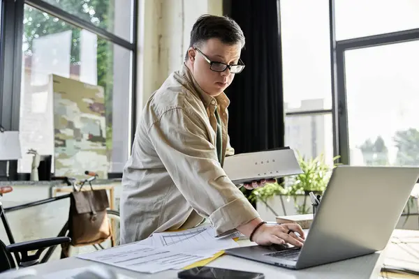 A young man with Down syndrome works on a laptop in a brightly lit office. — Stock Photo
