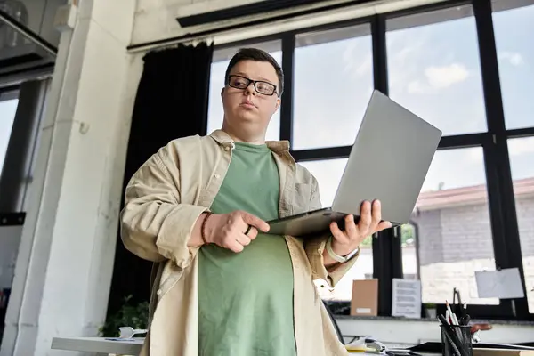 A young man with Down syndrome stands in a bright office, holding a laptop in his hands. — Stock Photo