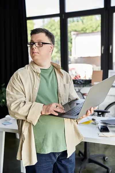 Um jovem com síndrome de Down, usando óculos, fica em um escritório usando um laptop. — Fotografia de Stock