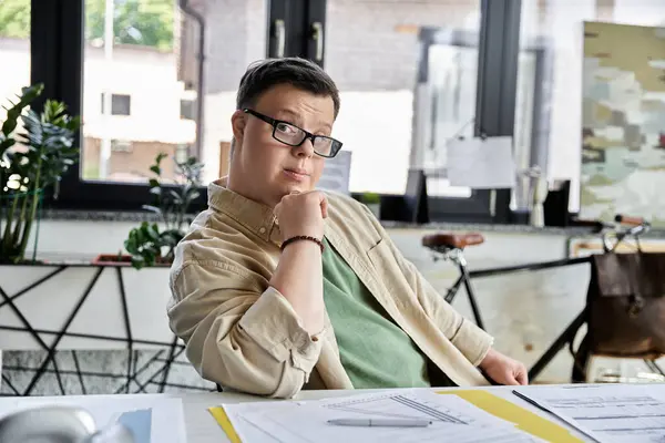 A young man with Down syndrome sits at a desk, thoughtfully resting his chin on his hand. — Stock Photo