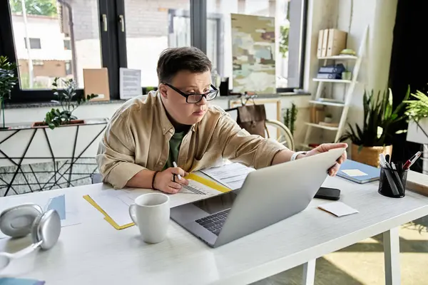 A young man with Down syndrome works on his laptop in a bright office. — Stock Photo
