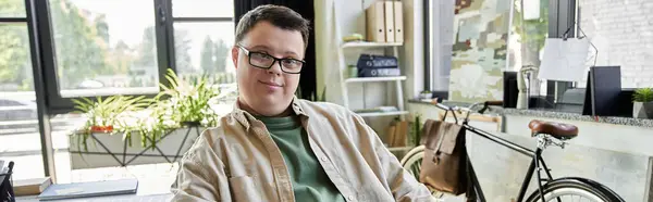 A young man with Down syndrome sits in a modern office with a bicycle in the background. — Stock Photo
