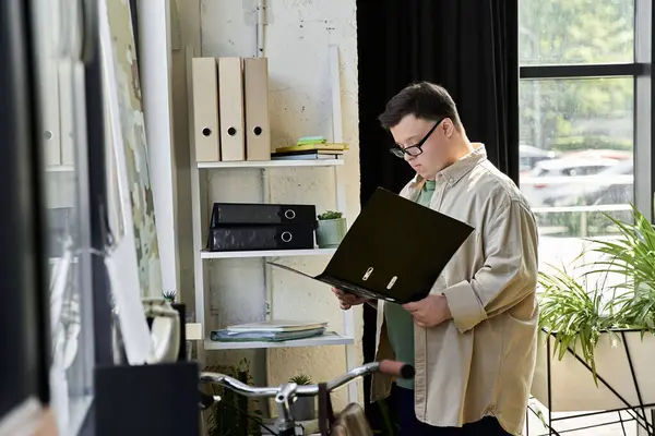 A young man with Down syndrome stands near a bookshelf, reviewing documents. — Stock Photo