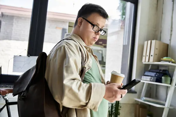 A young man with Down syndrome, wearing glasses, checks his phone while holding a cup of coffee. — Stock Photo