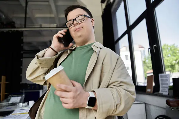 A young man with Down syndrome holds a coffee cup while talking on his phone. — Stock Photo