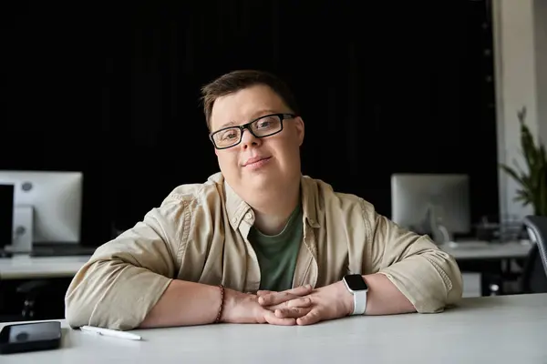 A young man with Down syndrome sits at a desk with a relaxed, confident smile. — Stock Photo
