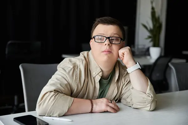 A young man with Down syndrome sits at a desk, resting his chin on his hand, contemplating. — Stock Photo