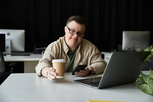 Ein junger Mann mit Down-Syndrom sitzt an einem Schreibtisch und blickt auf sein Handy, mit einer Tasse Kaffee in der anderen Hand.. — Stockfoto