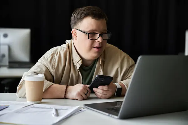 Ein junger Mann mit Down-Syndrom sitzt an einem Schreibtisch und arbeitet an seinem Laptop, während er ein Smartphone in der Hand hält. — Stockfoto