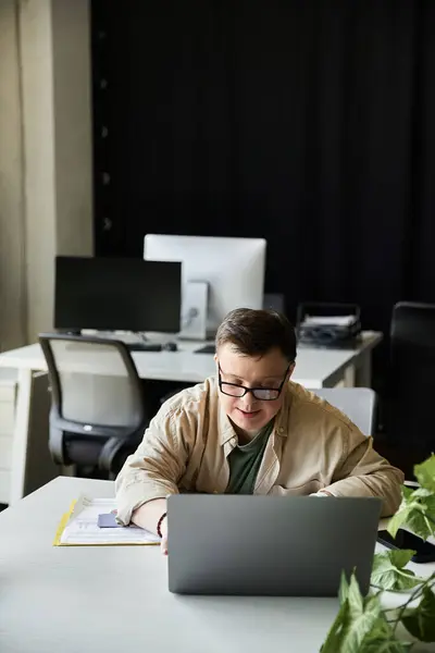 A young man with Down syndrome sits at a desk, working on a laptop. — Stock Photo