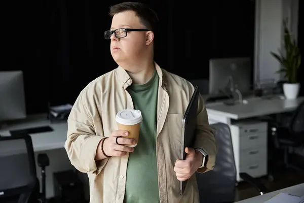 Ein junger Mann mit Down-Syndrom hält eine Tasse Kaffee und einen Laptop im Büro. — Stockfoto