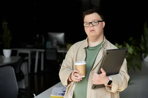 Un jeune homme trisomique se tient dans un espace de travail, tenant une tasse de café et un ordinateur portable. — Photo de stock