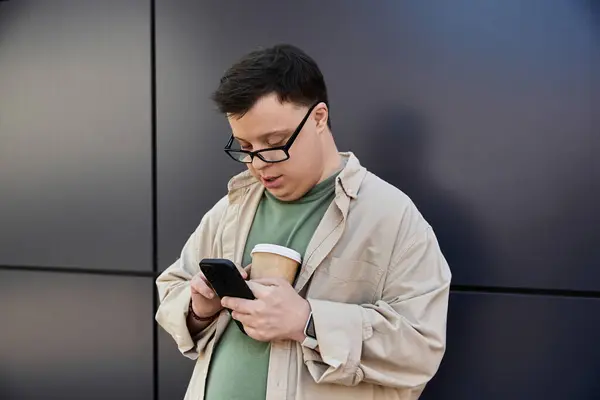 A man with Down syndrome stands by a wall, looking at his phone while holding a cup of coffee. — Stock Photo