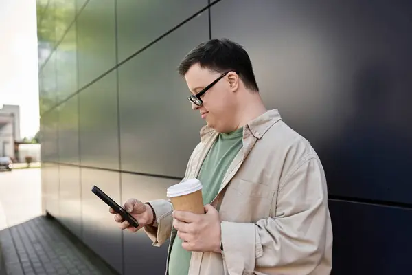 A man with Down syndrome stands outside, holding a cup of coffee and texting on his phone. — Stock Photo