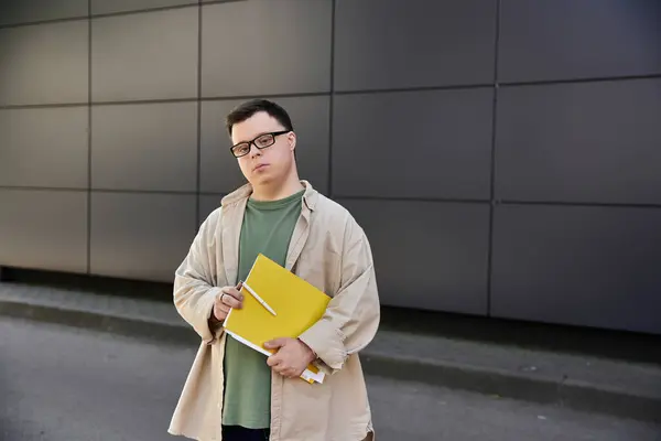 A man with Down syndrome holds books in front of a building in a city. — Stock Photo