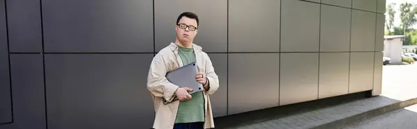A young man with Down syndrome stands outside, holding a laptop. — Stock Photo