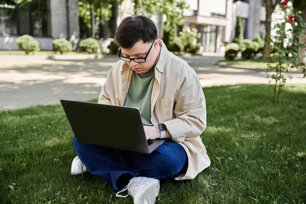 A young man with Down syndrome sits on the grass in a park, using a laptop. — Stock Photo