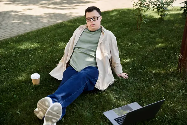 A young man with Down syndrome sits on the grass with a coffee cup and laptop. — Stock Photo