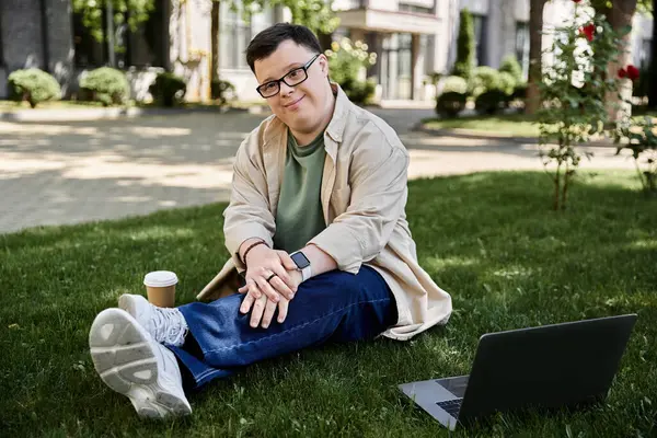 Ein junger Mann mit Down-Syndrom sitzt im Gras und genießt einen Kaffee und seinen Laptop. — Stockfoto