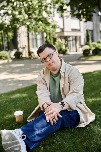 A young man with Down syndrome sits on the grass, looking thoughtful. — Stock Photo
