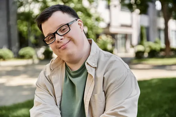 A smiling young man with Down syndrome in glasses sits in a park looking at the camera — Stock Photo