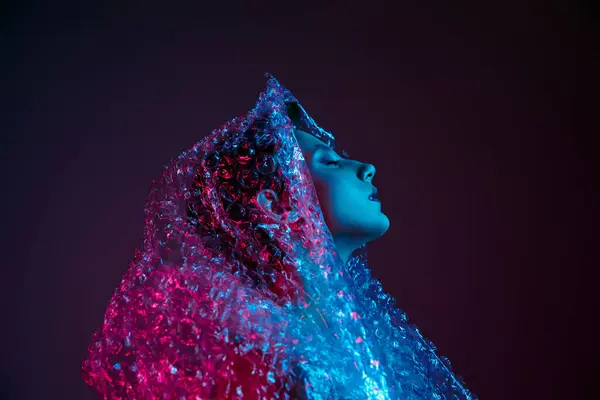 A woman enveloped in a shimmering blue and pink bubble wrap, eyes closed and lost in thought. — Stock Photo