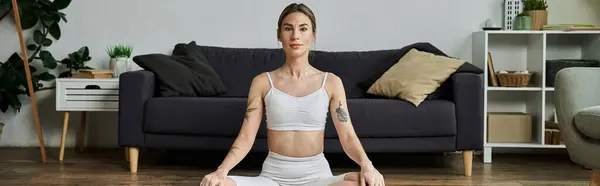 In a modern apartment, a young woman in active wear sits cross-legged on the floor practicing yoga, looking calm and focused — Stock Photo