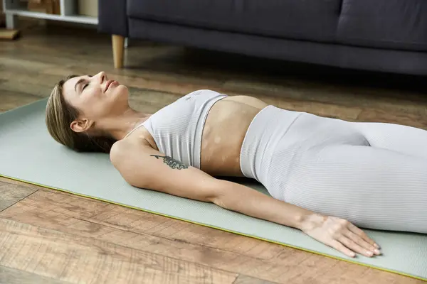 A young woman with vitiligo is lying on her back on a yoga mat in a modern apartment, eyes closed, practicing yoga or meditation. — Stock Photo