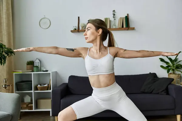 A young woman in white activewear performs a yoga pose in a modern living room. — Stock Photo