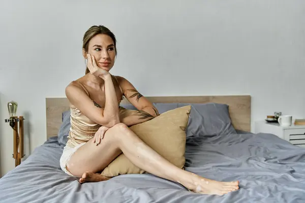 A young woman with vitiligo and a tattoo sits on a bed in a modern apartment, resting her chin on her hand and looking thoughtfully ahead. — Stock Photo