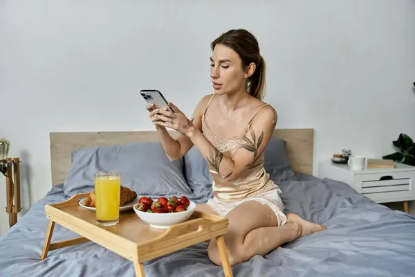 A young woman with vitiligo and a tattoo sits on her bed, enjoying a leisurely morning with a phone, breakfast, and a cozy atmosphere. — Stock Photo