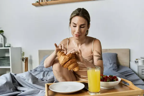 A woman in her 20s with vitiligo sits in bed, breaking a croissant in half. — Stock Photo