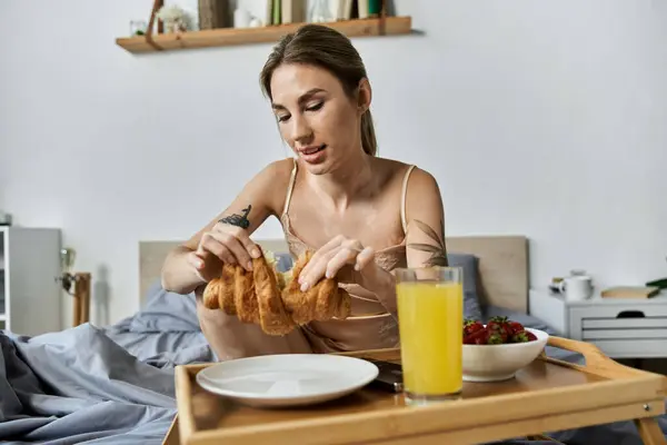 A young woman with vitiligo and a tattoo enjoys a croissant and orange juice while sitting on her bed in a modern apartment. — Stock Photo