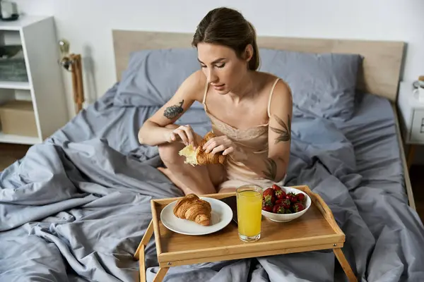 Eine Frau in ihren 20ern sitzt im Bett und genießt ein Frühstück mit Croissants und Orangensaft. — Stockfoto