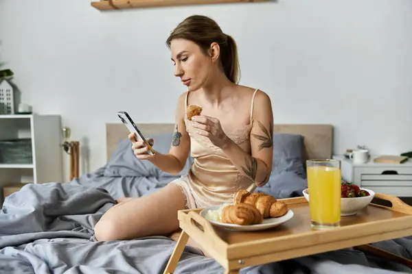 A young woman in her 20s enjoys breakfast in bed, checking her phone while eating croissants and drinking juice. — Stock Photo