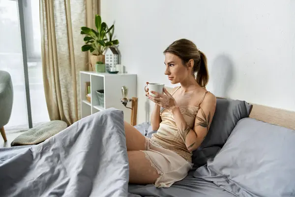 A young woman with vitiligo and a tattoo sits in bed, enjoying a cup of coffee in her modern apartment. — Stock Photo
