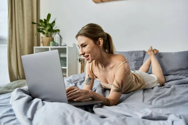 A woman with vitiligo and a tattoo smiles as she works on her laptop in bed, surrounded by cozy blankets and a plant. — Stock Photo