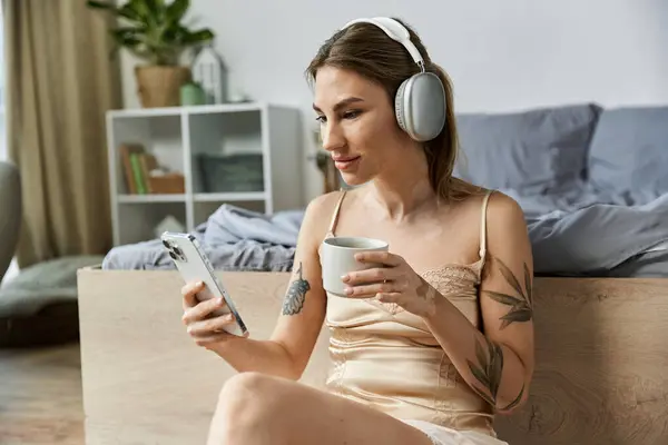 A young woman with vitiligo and a tattoo sits on the edge of her bed, enjoying a cup of coffee while scrolling on her phone. — Stock Photo