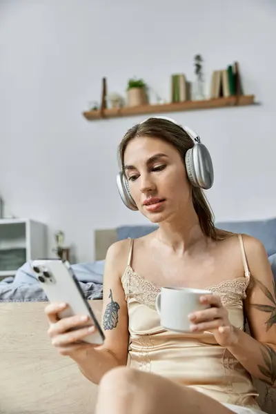 A young woman with vitiligo relaxes in her modern apartment bedroom, enjoying a cup of coffee and browsing her phone. — Stock Photo
