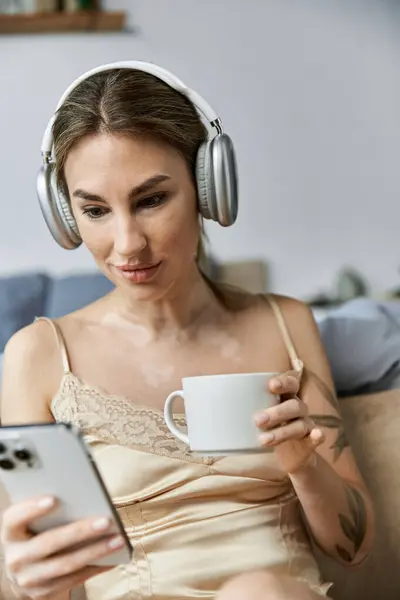 Sulla ventina, una donna con la vitiligine e un tatuaggio si rilassa nella sua camera da letto moderna con caffè, scorrendo sul telefono e indossando cuffie — Foto stock