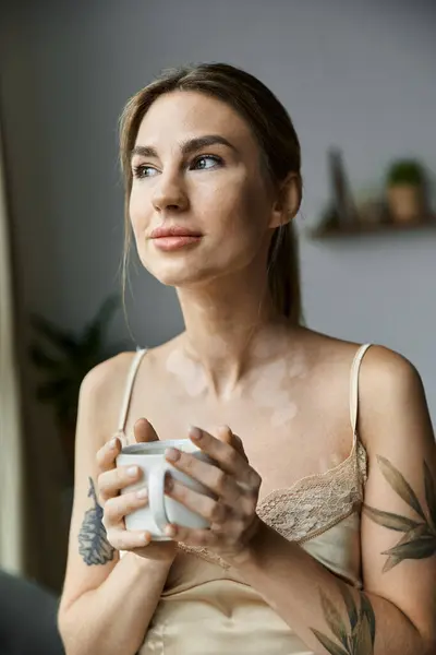A young woman with vitiligo and a tattoo, sits in her bedroom, holding a mug and gazing out a window. — Stock Photo