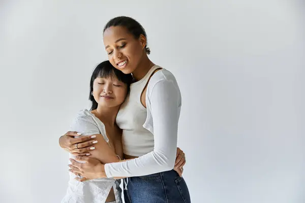 Due giovani donne, una afroamericana e una asiatica, si abbracciano su un semplice sfondo grigio. — Foto stock