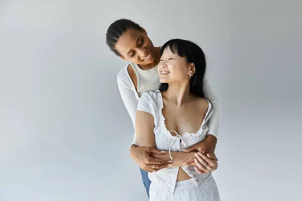 Una mujer afroamericana sostiene a una mujer asiática cerca mientras comparten un abrazo amoroso contra un fondo gris. — Stock Photo