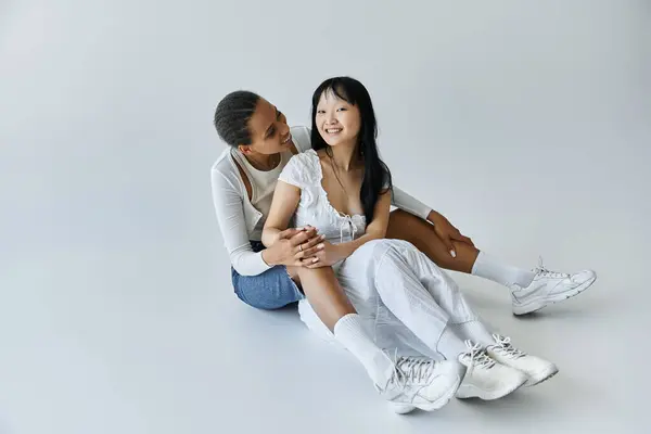 Two young women, one Black and one Asian, sit on a grey background, holding hands and smiling at each other. — Stock Photo